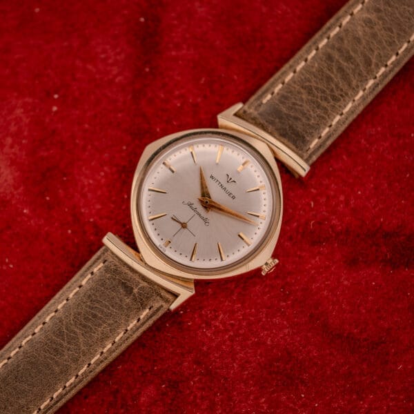 The Wittnauer Automatic Ref. 2598-2 with Cal. 11AO, featuring an asymmetric case and brown leather strap, rests elegantly on a red textured surface.