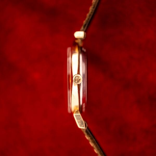 Side view of a Wittnauer Automatic Ref. 2598-2 Asymmetric Case gold wristwatch with Cal. 11AO movement and brown strap, displayed on a textured red background.