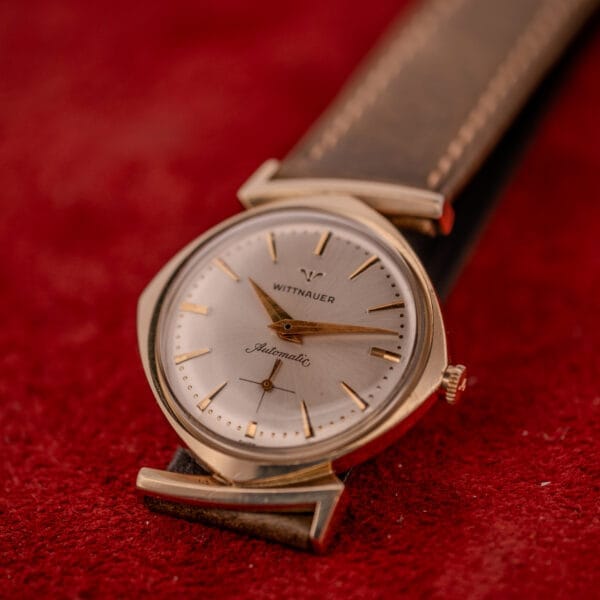 A Wittnauer Automatic Ref. 2598-2 with an asymmetric case and brown leather strap rests on a red textured surface.