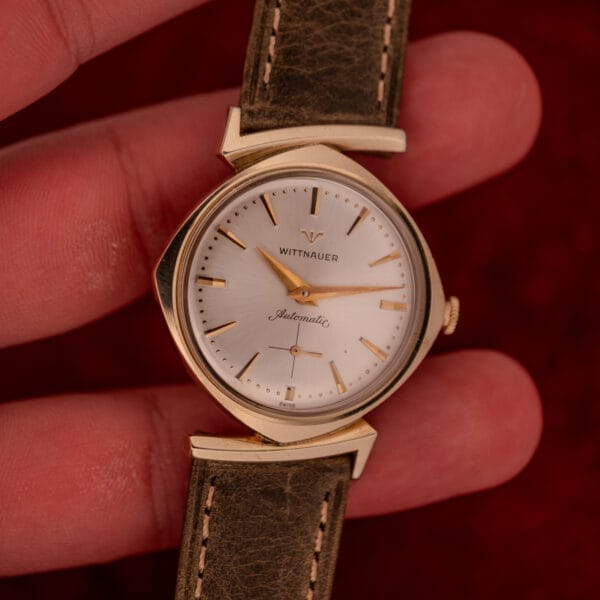 A hand holding a Wittnauer Automatic Ref. 2598-2 wristwatch featuring an asymmetric case, Cal. 11AO movement, brown leather strap, and gold finish.