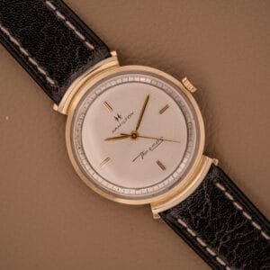 A Hamilton Thin-O-Matic Ref. T-406 with hooded lugs, Micro Rotor Cal. 663, and black leather strap is displayed on a tan surface.