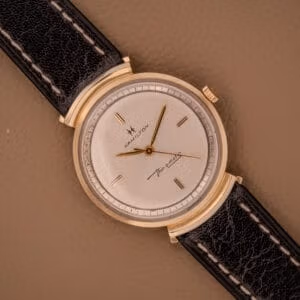 A Hamilton Thin-O-Matic Ref. T-406 with hooded lugs, Micro Rotor Cal. 663, and black leather strap is displayed on a tan surface.