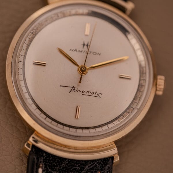 Close-up of a Hamilton Thin-O-Matic Ref. T-406, showcasing hooded lugs, a gold case, micro rotor Cal. 663 movement, and a black leather strap.