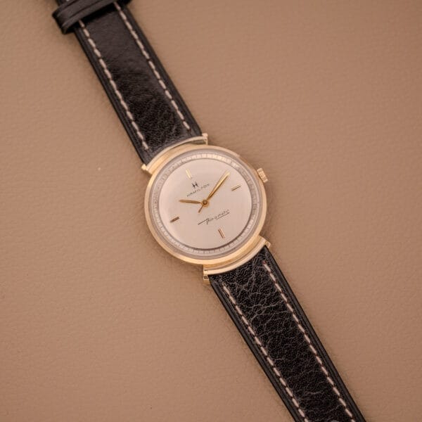 A Hamilton Thin-O-Matic Ref. T-406 with hooded lugs and a black leather strap, featuring a micro rotor Cal. 663 movement, rests on a beige surface.