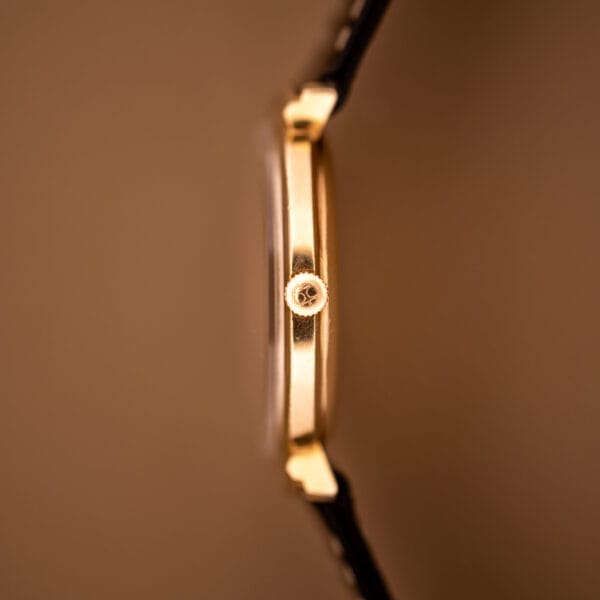 Side view of a Hamilton Thin-O-Matic Ref. T-406 Hooded Lugs wristwatch with black strap, visible crown, slim profile, and Micro Rotor Cal. 663 movement, set against a brown background.