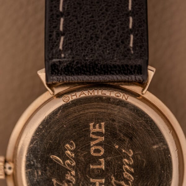 Close-up of a gold Hamilton Thin-O-Matic Ref. T-406 watch case back with engraved text, featuring hooded lugs, micro rotor Cal. 663 movement, and a black leather strap.