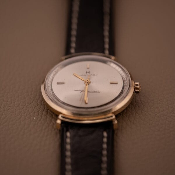 A Hamilton Thin-O-Matic Ref. T-406 Hooded Lugs Micro Rotor Cal. 663 wristwatch with a black leather band and gold casing is displayed on a brown textured surface.