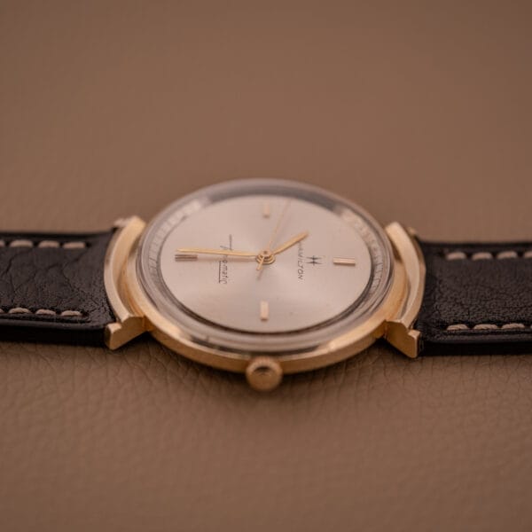 A Hamilton Thin-O-Matic Ref. T-406 with hooded lugs and Cal. 663 Micro Rotor movement, paired with a black leather strap, is elegantly displayed on a brown surface.