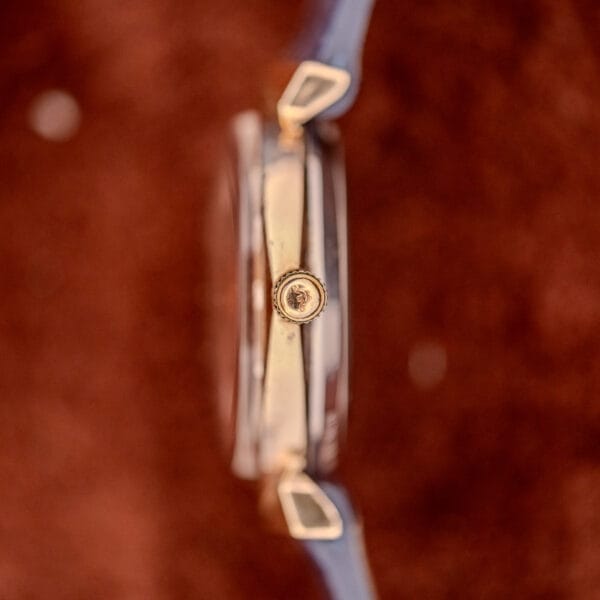 Side view of a Wittnauer Automatic Ref. 2598-1 Hooded Lugs Cal. 11SR wristwatch with a gold case, blue strap, set against a textured brown background.