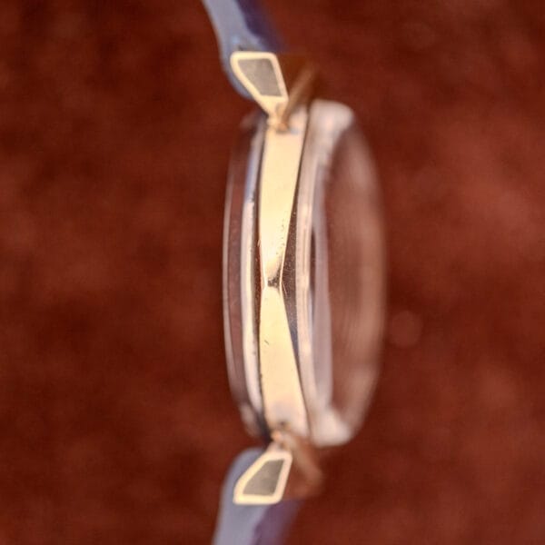 Side view of a Wittnauer Automatic Ref. 2598-1 Hooded Lugs Cal. 11SR gold and clear case wristwatch with a blue strap, shown against a brown background.