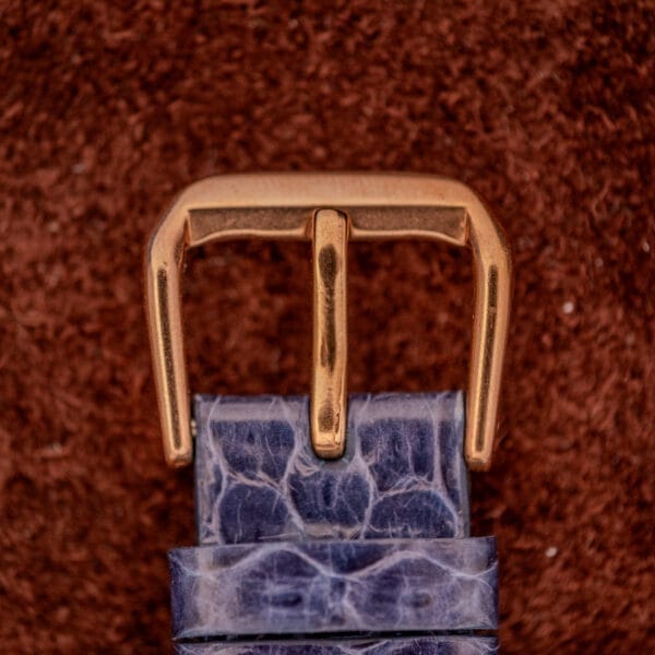 A gold belt buckle on a textured blue leather strap, set against a brown background, evokes the refined style of the Wittnauer Automatic Ref. 2598-1 Hooded Lugs Cal. 11SR.