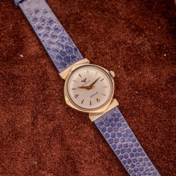 The Wittnauer Automatic Ref. 2598-1 Hooded Lugs Cal. 11SR, with a gold case, silver dial, and blue textured leather strap, is elegantly displayed on a brown textured surface.