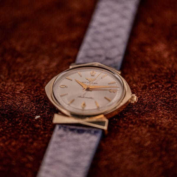 A gold Wittnauer Automatic Ref. 2598-1 Hooded Lugs Cal. 11SR wristwatch with a gray textured strap rests elegantly on a brown suede surface.