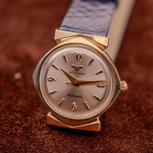 The Wittnauer Automatic Ref. 2598-1 Hooded Lugs Cal. 11SR wristwatch features a silver dial and blue leather strap, showcased on a brown textured surface.