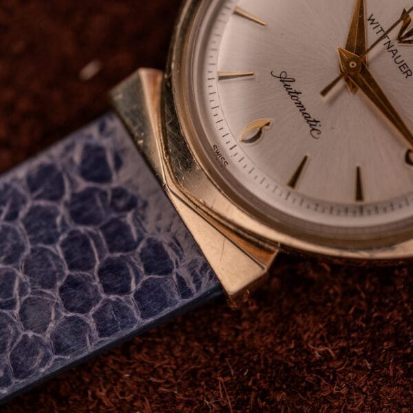 Close-up of a Wittnauer Automatic Ref. 2598-1 Hooded Lugs Cal. 11SR wristwatch with a gold case and textured blue leather strap, displayed on a brown surface.
