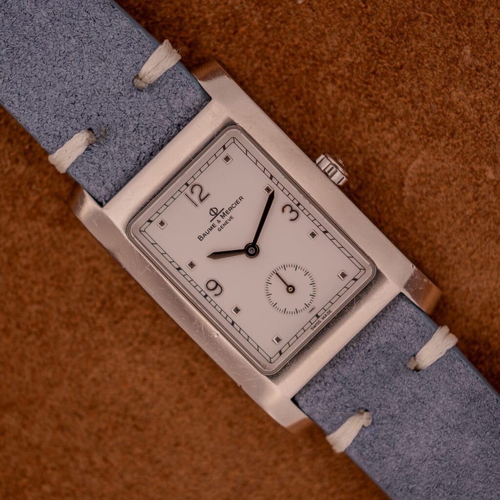 Baume Mercier Hampton Large MV05063 wristwatch with a white dial and light blue leather strap, shown on a brown textured surface.
