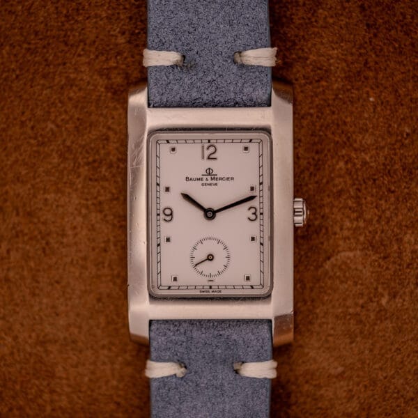 A Baume Mercier Hampton Large MV05063 Quartz watch with a white dial and blue strap is displayed on a brown textured surface.