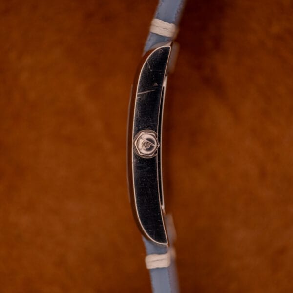 Side view of the Baume Mercier Hampton Large MV05063 wristwatch with a blue strap, white dial quartz movement, set against a textured brown background.