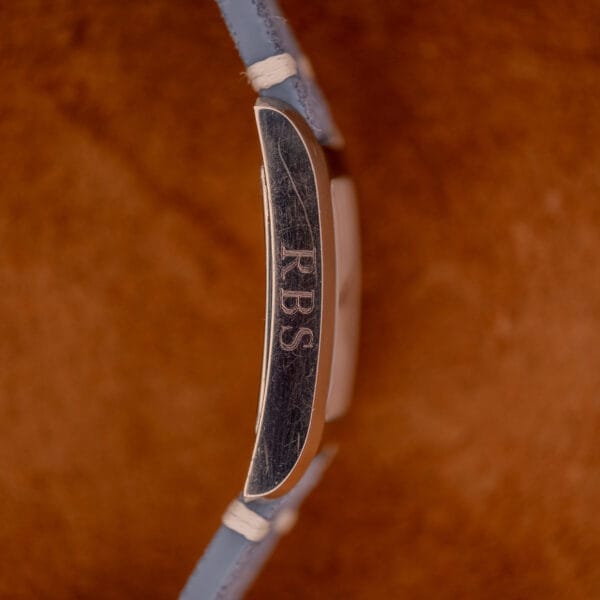 Close-up of a metal object engraved with RBS, held by light blue straps, evoking the elegance of the Baume Mercier Hampton Large MV05063 White Dial Quartz watch, set against a brown background.