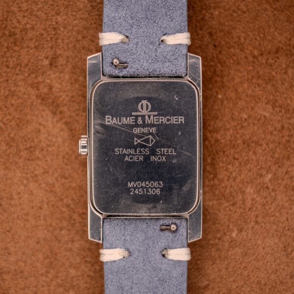 Back of a Baume Mercier Hampton Large MV05063 White Dial Quartz wristwatch with engravings and a grey suede strap, displayed on a brown background.