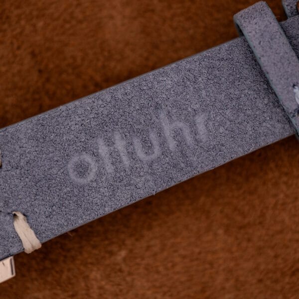 Close-up of a grey suede strap with "otuhr" embossed, set against a brown background, evoking the refined aesthetic of the Baume Mercier Hampton Large MV05063 White Dial Quartz.