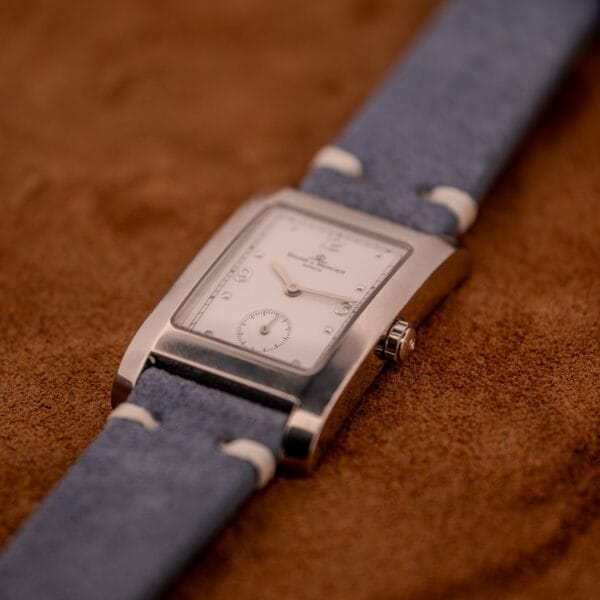 A Baume Mercier Hampton Large MV05063 white dial quartz wristwatch with a blue suede strap rests on a brown suede surface.