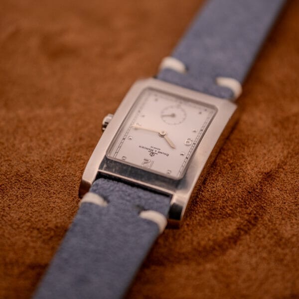 The Baume Mercier Hampton Large MV05063 White Dial Quartz wristwatch with a gray suede strap is displayed on a brown suede surface.