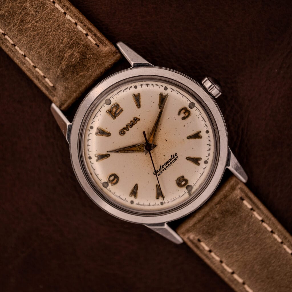 A Cyma Automatic Watersport Bumper 2-1221 Stainless Steel Cal. R420 wristwatch with a beige dial, brown numerals, and a tan leather strap showcased on a brown background.