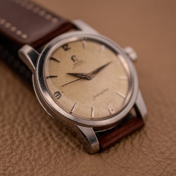 An Omega Automatic Seamaster Ref. 2846-2848 Stainless Steel Cal. 500 watch with a cream dial and brown leather strap rests on a tan surface.