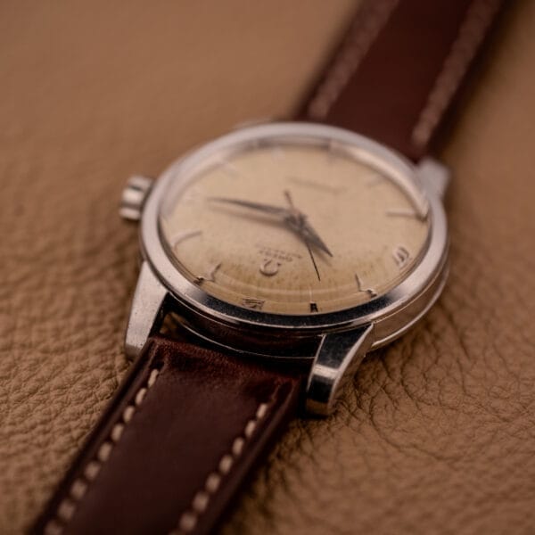 Close-up of an Omega Automatic Seamaster Ref. 2846-2848 Stainless Steel Cal. 500 vintage watch with a brown leather strap, displayed on a textured tan surface.