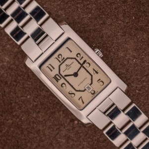 The Baume Mercier Hampton Automatic Large MV045120 features a rectangular stainless steel case, beige dial, and link band, presented on a brown background.