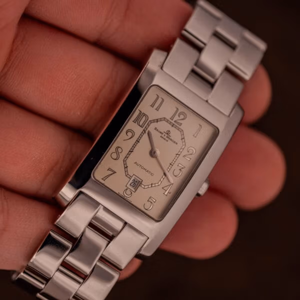 Baume Mercier Hampton MV045120 held in hand at angle, full bracelet and rectangular case visible, dial showing AUTOMATIC text and date window.