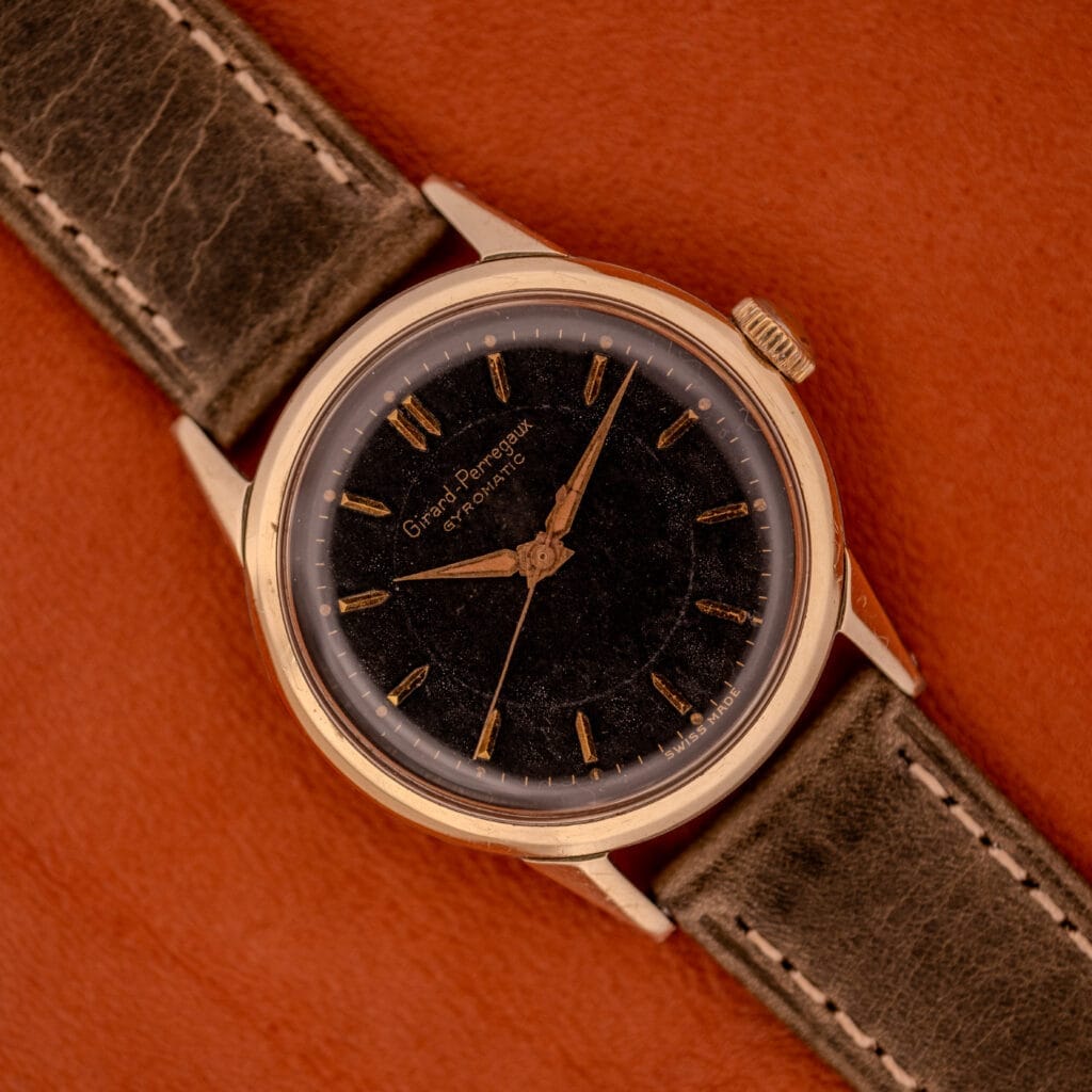 A Girard Perregaux Gyromatic Ref. 6250 wristwatch with a black gilt dial and gold capped case is showcased with a brown leather strap on a brown surface.