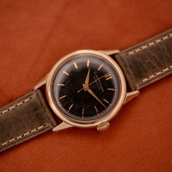 A Girard Perregaux Gyromatic Black Gilt Dial Ref. 6250 with a gold capped case and worn leather strap rests elegantly on a brown leather surface.