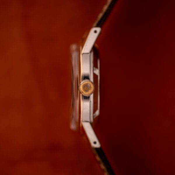 Side view of a Girard Perregaux Gyromatic Black Gilt Dial Ref. 6250 with a gold capped case and round crown, set against a blurred reddish-brown background.