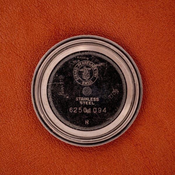 Round metal case back of the Girard Perregaux Gyromatic Black Gilt Dial Ref. 6250 Gold Capped Case on orange textured surface, engraved with STAINLESS STEEL 6250 M 194 R—an iconic detail on this vintage model.