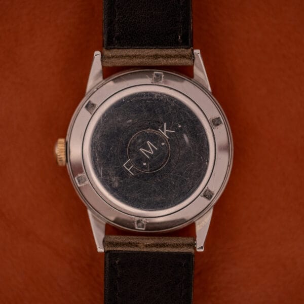 A Girard Perregaux Gyromatic Black Gilt Dial Ref. 6250 with a gold capped case, F.M.K engraved on the back, and a black leather strap rests elegantly on a brown surface.