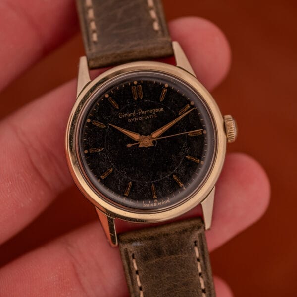 A hand holding a Girard Perregaux Gyromatic Black Gilt Dial Ref. 6250, featuring a gold capped case and brown leather strap.