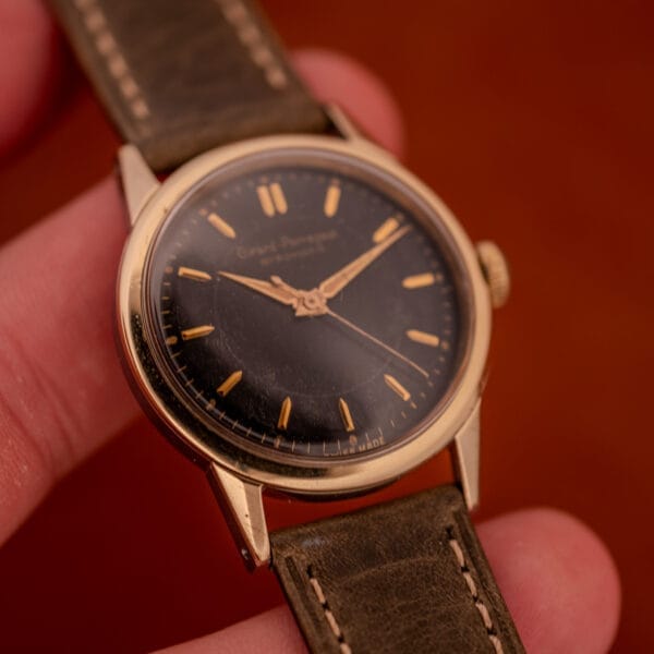 A hand holds a Girard Perregaux Gyromatic Black Gilt Dial Ref. 6250 with a gold capped case and brown leather strap against a brown background.