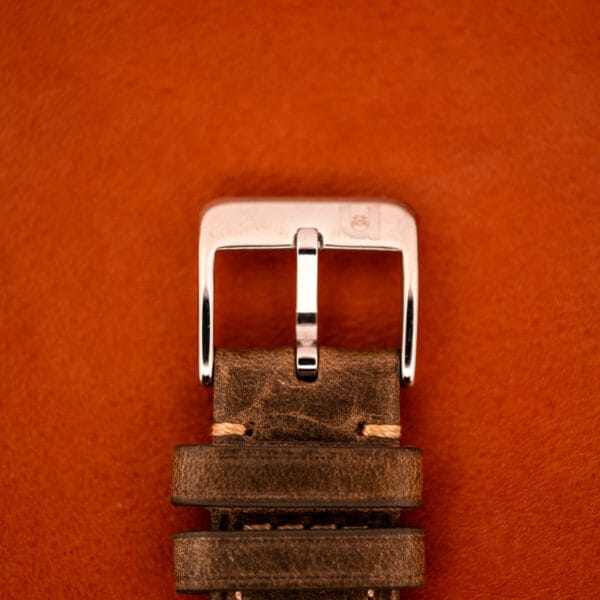 A close-up of a brown leather watch strap with a silver buckle on an orange background, paired perfectly with the Girard Perregaux Gyromatic Black Gilt Dial Ref. 6250 Gold Capped Case.