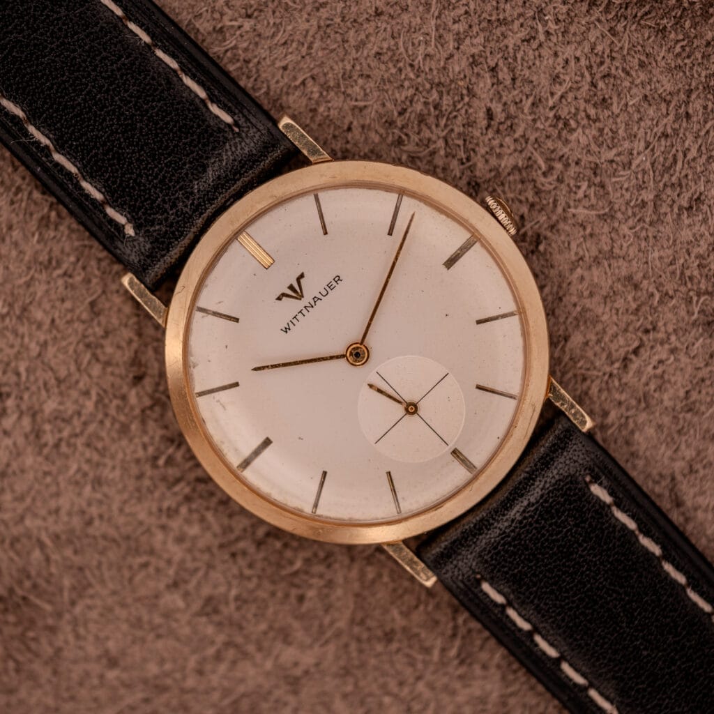 A Wittnauer Ref. 2028 Manual Wind Cal. 11BG2 wristwatch with a white dial and gold indexes rests elegantly on a textured brown surface.
