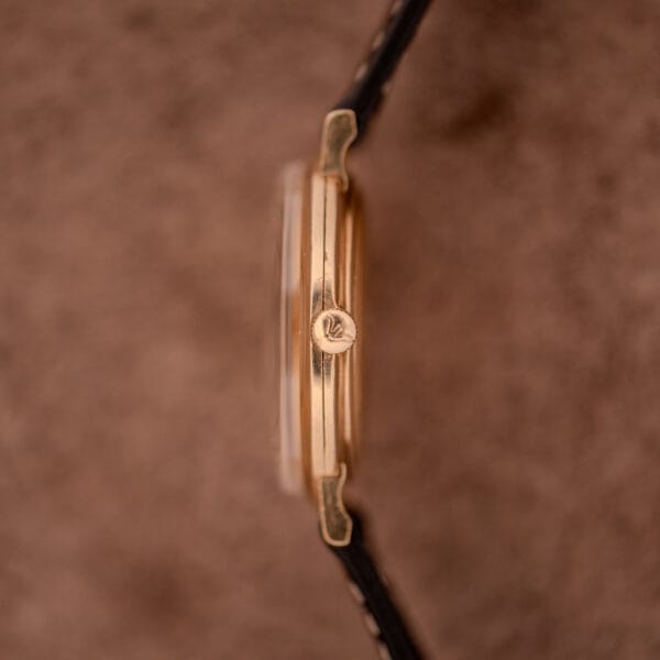 Side view of a Wittnauer Ref. 2028 Manual Wind Cal. 11BG2 wristwatch with a white dial, gold indexes, and black strap, shown against a soft brown background.