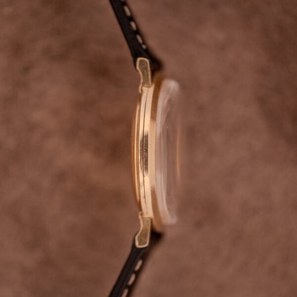 Side view of a Wittnauer Ref. 2028 Manual Wind Cal. 11BG2 wristwatch with a white dial, gold indexes, and black strap, set against a blurred brown background.