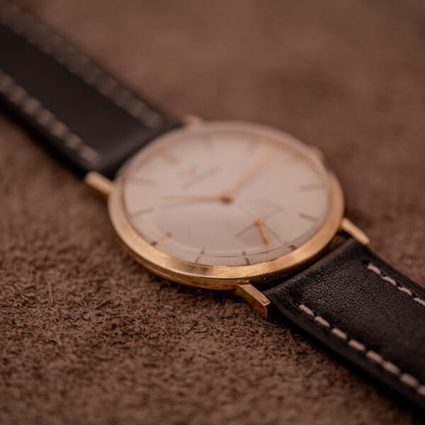 Wittnauer Ref. 2028 Manual Wind Cal. 11BG2 wristwatch with a white dial and gold indexes, shown close-up on a brown textured surface with a black leather strap.