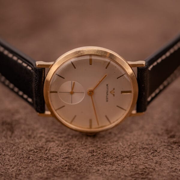 Wittnauer Ref. 2028 manual wind watch with Cal. 11BG2 movement, white dial, gold indexes, and black leather strap rests on a brown textured surface at 10:10.