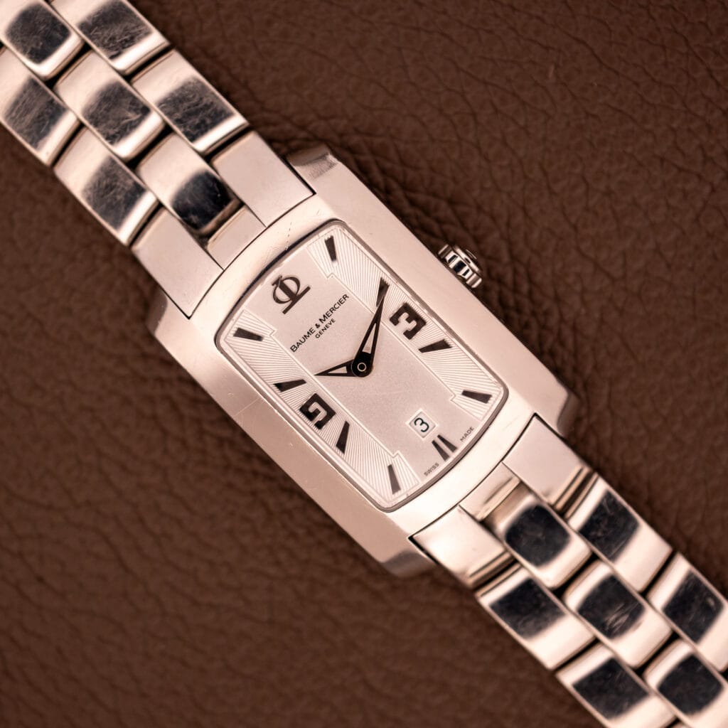 Baume Mercier Hampton 65511 Quartz Stainless Steel wristwatch with rectangular silver case, shown on a textured brown leather background.