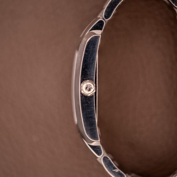 A close-up side view of the Baume Mercier Hampton 65511 Quartz Stainless Steel watch case and crown set against a brown background.