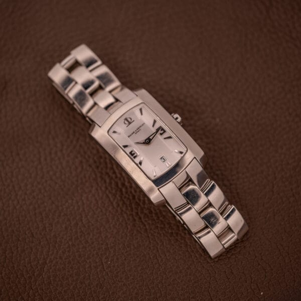 A Baume Mercier Hampton 65511 Quartz Stainless Steel wristwatch, featuring a silver rectangular case and band, rests on a brown textured surface.