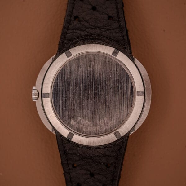 A close-up of a scratched silver Omega Automatic Geneve Dynamic Ref. 166.107 Cal. 1421 watch back with a black leather strap on a brown background.