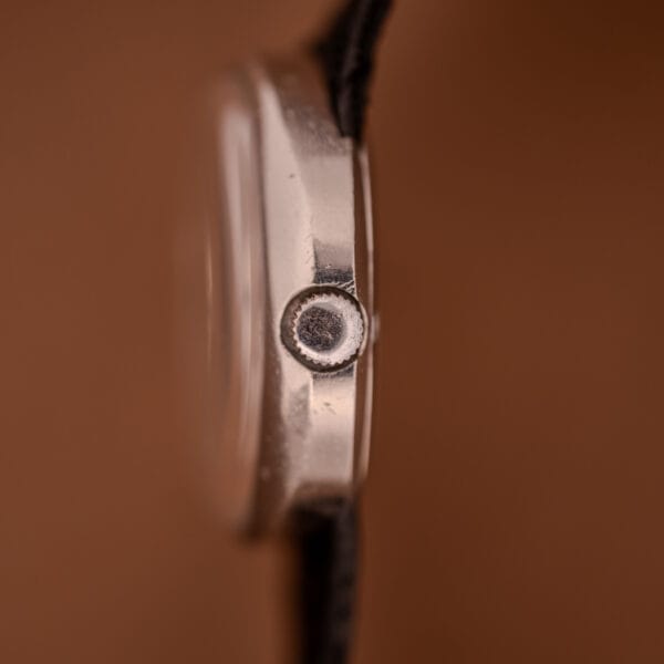 Side view of an Omega Automatic Geneve Dynamic Ref. 166.107 Cal. 1421 wristwatch crown in silver with a black strap, set against a brown background.